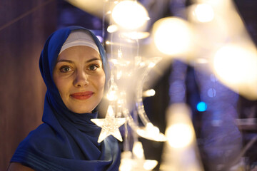 Fototapeta premium Smiling Muslim Woman In Blue Hijab Among Star And Moon String Lights During Festive Evening