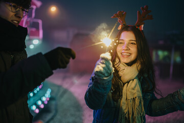 Fototapeta premium Two friends hold a lit Christmas sparkler at night, one wearing reindeer antlers. A smiling woman in a scarf enjoys a festive sparkler outdoors with a friend.