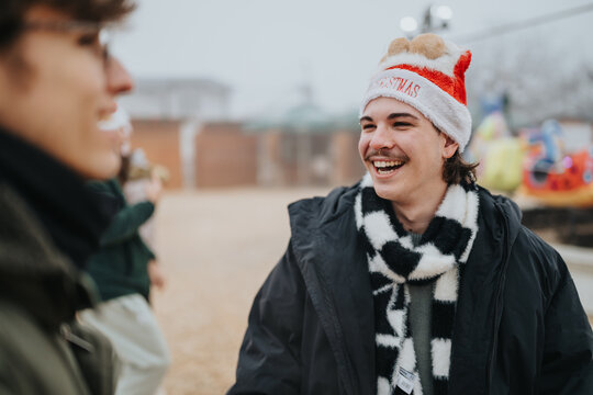 A young man wearing a Santa hat smiles and laughs while talking with friends outdoors. The festive scene shows casual winter clothing and a cheerful atmosphere near an amusement ride.