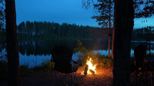 Bright campfire burning at a campsite between dark trees on a lake shore during blue hour twilight. Concept of night wild camping, romantic outdoor recreation, peaceful wilderness getaway and twilight