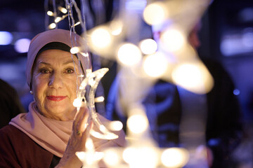 Fototapeta premium Elderly Woman In Hijab Holding Crescent Light Decoration At Festive Night Market Celebration