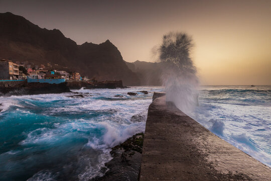 View of a large wave crashing against a concrete pier at sunset with the rugged coastline and buildings of Santo Antao in the background, Ponta do Sol, Ribeira Grande, Cabo Verde.