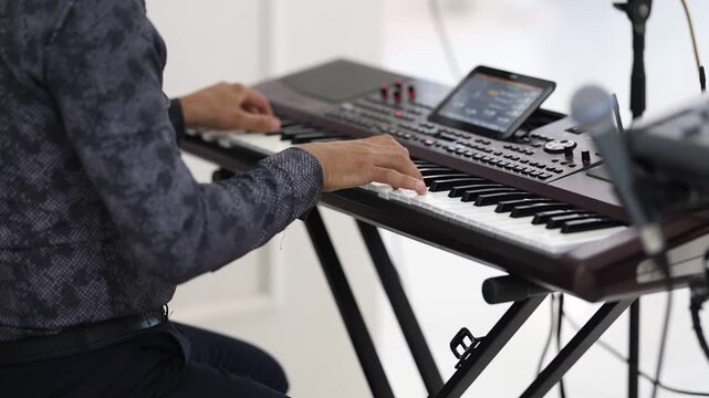 Close-up musician hands playing electronic keyboard workstation in bright studio, with right hand pressing white and black keys while left hand reaches toward upper keys and tilted control screen