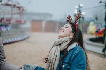 Fototapeta premium A young woman wearing a reindeer antler headband laughs while bundled in a scarf and denim jacket. She enjoys a festive moment at an outdoor winter amusement park or holiday market.