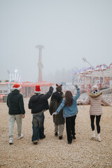 Fototapeta premium A group of friends wearing winter coats and Santa hats walk toward a misty amusement park. The festive group strolls across gravel toward rides and lights in a foggy winter scene.