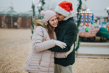 Fototapeta premium A young couple hugs and smiles at an outdoor winter fair while wearing warm coats and a Santa hat. They share a festive moment together against blurred amusement rides and holiday lights.