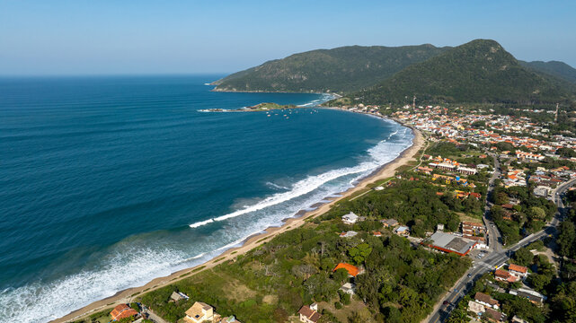 Aerial view of the coastline of Florianopolis, featuring sandy beaches, turquoise ocean waves, lush green hills, and residential buildings in Florianopolis, Santa Catarina, Brazil.