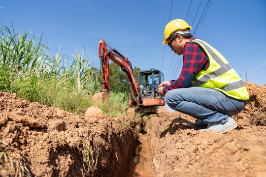 An Engineer using a tablet to inspect a trench prepared for water pipe installation with an excavator, representing civil engineering, utilities and water infrastructure.