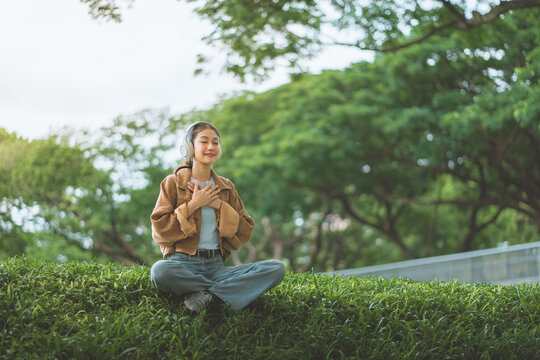 Young woman wearing headphones practicing mindfulness with hands on heart while relaxing in green nature