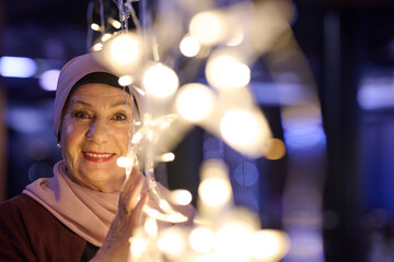 Fototapeta premium Elderly Woman Wearing Hijab Smiling With Festive String Lights And Warm Bokeh Portrait