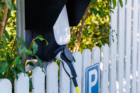 Close up shot of electric vehicle charging station wallbox with plugged cable next to white fence and green foliage, EV technology and sustainable mobility concept