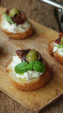 Vertical video. Cooking bruschetta or crostini. Pouring olive oil over toasted bread with cream cheese, sun dried tomatoes and olives.