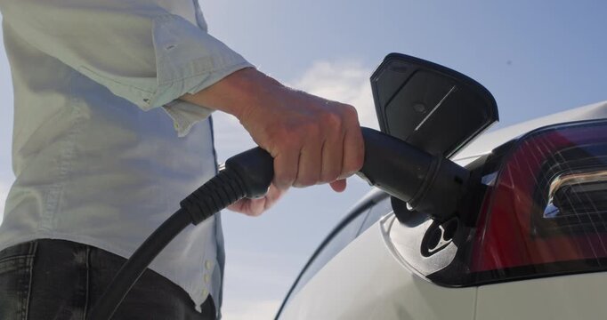 Man, in the rays of the backlight, connects his electric car to a charging station, inserting the plug into the socket of the opened charging hatch.