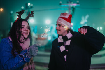 Fototapeta premium Two friends celebrate Christmas outdoors at night holding lit sparklers and smiling. A festive moment shows them wearing a Santa hat, reindeer antlers and warm winter clothing.