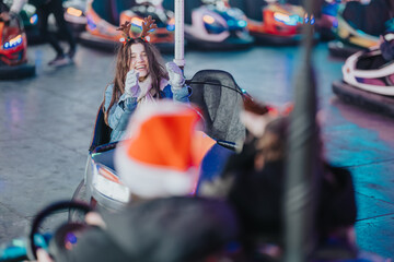 Fototapeta premium A teenage girl wearing reindeer antlers laughs while riding in a bumper car at a holiday fair. The colorful, festive scene shows joyful amusement park fun with lights and seasonal costumes.