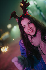 Fototapeta premium A young woman wearing reindeer antlers smiles while holding a sparkler at night. A festive nighttime portrait showing holiday cheer among friends.
