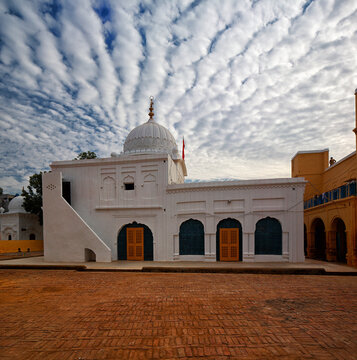 View of the white Gurudwara Patti Sahib with its distinctive dome and arched doorways set against a dramatic cloudy sky in the brick courtyard of Nankana Sahib, Punjab, Pakistan.