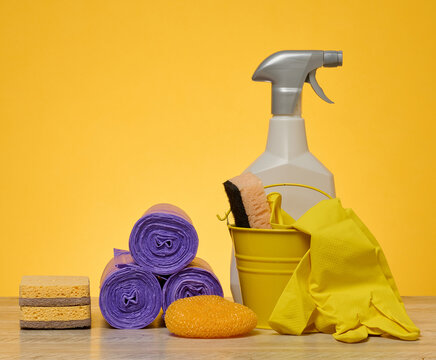 Cleaning set consisting of yellow bucket, rubber gloves, sponges, purple garbage bags and spray bottle on wooden table