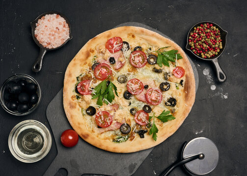 Whole pizza on a wooden board surrounded by tomatoes, olives, herbs, spices and kitchen tools on a dark background