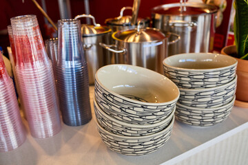 Fototapeta premium Stacks of patterned ceramic bowls and colorful plastic cups on white counter with stainless steel pots in background at Vietnamese street food cafe or restaurant.