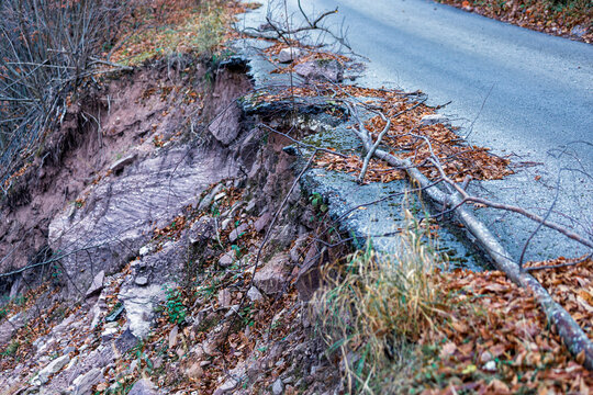 View of a damaged asphalt road with a significant landslide and erosion on the side, featuring fallen branches and autumn leaves on the rocky terrain in Topli Do, Pirot District, Serbia.