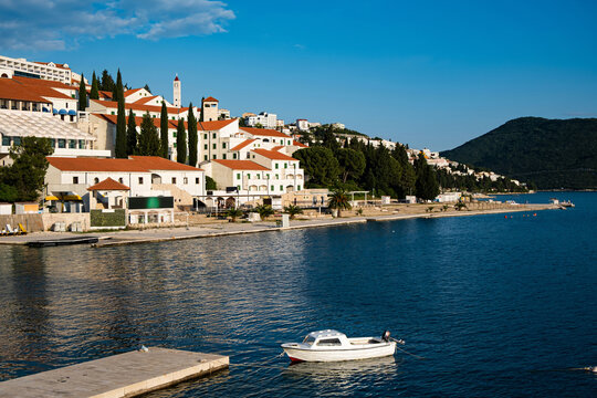 Coastal panorama of Neum, Bosnia and Herzegovina, with white waterfront buildings, red tile roofs, church tower, harbor, moored boat and wooded hills