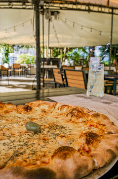 Margherita pizza with browned cheese, herbs, and olive on a plate beside water in an outdoor restaurant patio with string lights