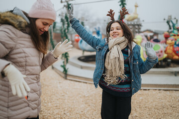 Fototapeta premium Two young women laugh and play outdoors wearing winter coats and reindeer antlers. The friends celebrate holiday fun together at a festive amusement ride in cold weather.