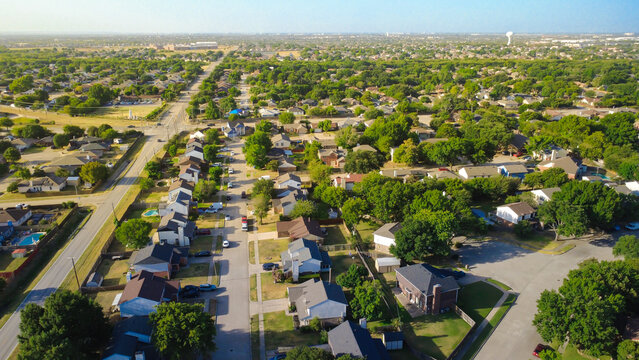 Long residential avenue extends past neat driveways toward distant Sublett Road commercial zones. Packed single family houses line concrete sidewalks, green lawns, sunny foliage, Arlington, Texas