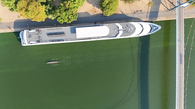 Aerial view of a cruise ship docked along the riverbank with a rowing boat moving through the green water in Frankfurt am Main, Hessen, Germany.