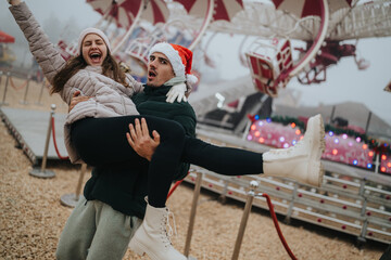 Fototapeta premium A young couple celebrate at a Christmas carnival with the man lifting the laughing woman. The festive scene shows joyful holiday fun at an outdoor amusement ride in winter.
