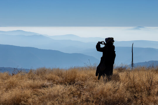 View of a person silhouetted against the hazy mountain layers of Stara planina while capturing a photo with a smartphone in the dry grass of Babin zub, Pirot District, Serbia.