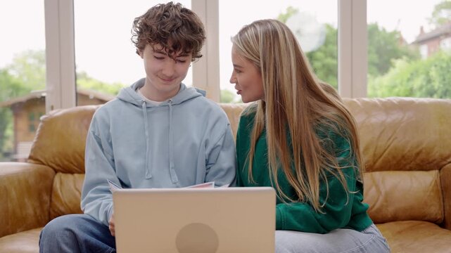 Mother and teenager boy sitting on a couch in a living room. They look at papers and a laptop. They smile and discuss homework. A piggy bank sits on the table near them.