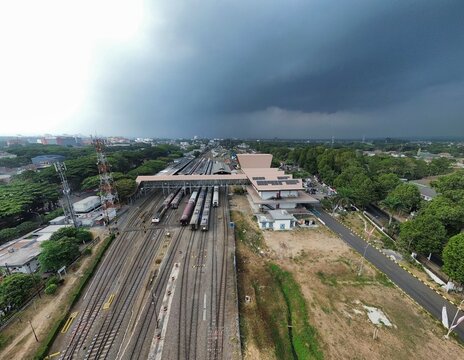 Aerial view of the Blimbing railway station with multiple train tracks, a pedestrian bridge, and surrounding lush greenery under a cloudy sky in Blimbing, East Java, Indonesia.