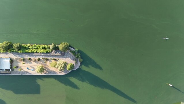 Aerial view of Westhafen Mole with lush trees, paved walkways, and long shadows stretching over the green water of the Main river in Frankfurt am Main, Hessen, Germany.
