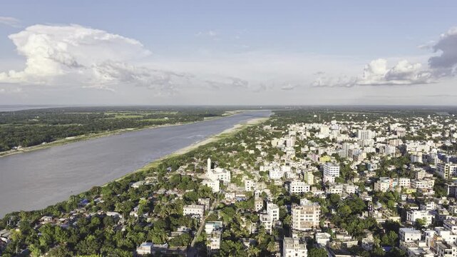 Aerial view of Kushtia, Bangladesh, featuring city buildings, the Gorai River along one side, open green landscape, and a blue cloudy sky during a forward drone movement.