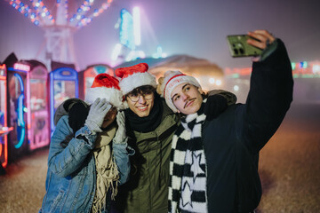 Fototapeta premium Three young friends wearing Santa hats smile for a selfie at night. They pose together in front of colorful carnival lights and rides.