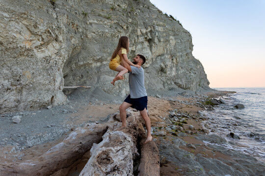 Happy father playing with daughter on a driftwood log by the rocky seaside cliffs at sunset, creating joyful family memories on vacation.