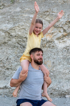Smiling father carries young daughter on shoulders outdoors, both looking happy and relaxed in summer clothing against rocky background.