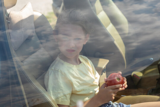 Young girl sitting inside a car, holding an apple, seen through a window with reflections of clouds and sky.
