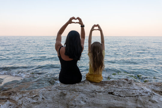 Back view of a woman and child sitting on driftwood at the beach, making heart shapes with their hands at sunset.
