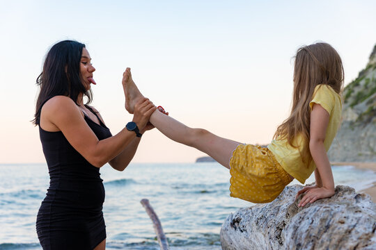 Playful mother and daughter enjoying time together by the sea, smiling and making faces at sunset on a rocky beach.