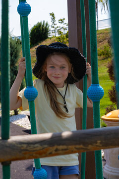 Cheerful young girl in a sun hat plays on outdoor playground ropes, enjoying a sunny day with green park scenery in the background.