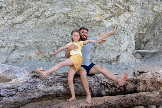 Happy father and daughter balancing on driftwood, enjoying a fun moment together against a rocky outdoor backdrop.