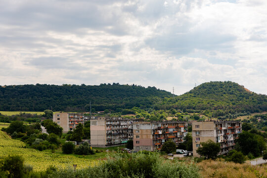 A cluster of aged apartment blocks surrounded by lush greenery and rolling hills under a cloudy sky, conveying rustic urban living.