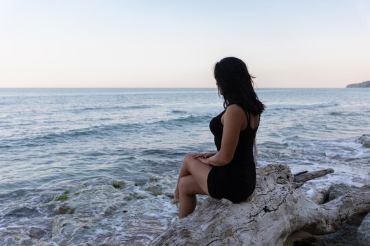 Woman in black dress sitting on driftwood, gazing at calm ocean waves under clear sky. Relaxed coastal scene with tranquil, natural atmosphere.