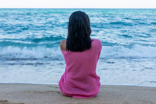 Back view of a woman in pink sitting on sandy beach facing calm blue ocean waves under natural daylight.