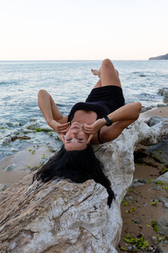 Confident woman in black dress lying on driftwood by the sea, serene beach setting with calm waves and clear sky, perfect for lifestyle or travel concepts.