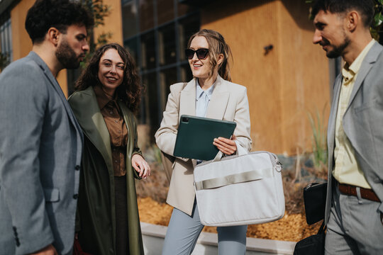 Four business colleagues talk outside while a woman holds a tablet and a laptop bag. Casual outdoor work meeting with professionals networking and sharing ideas.