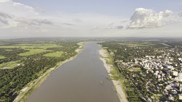 A forward-moving aerial view along the river shows the contrast between Kushtia's urban landscape on one side and green natural surroundings on the other, with distant clouds above the horizon.